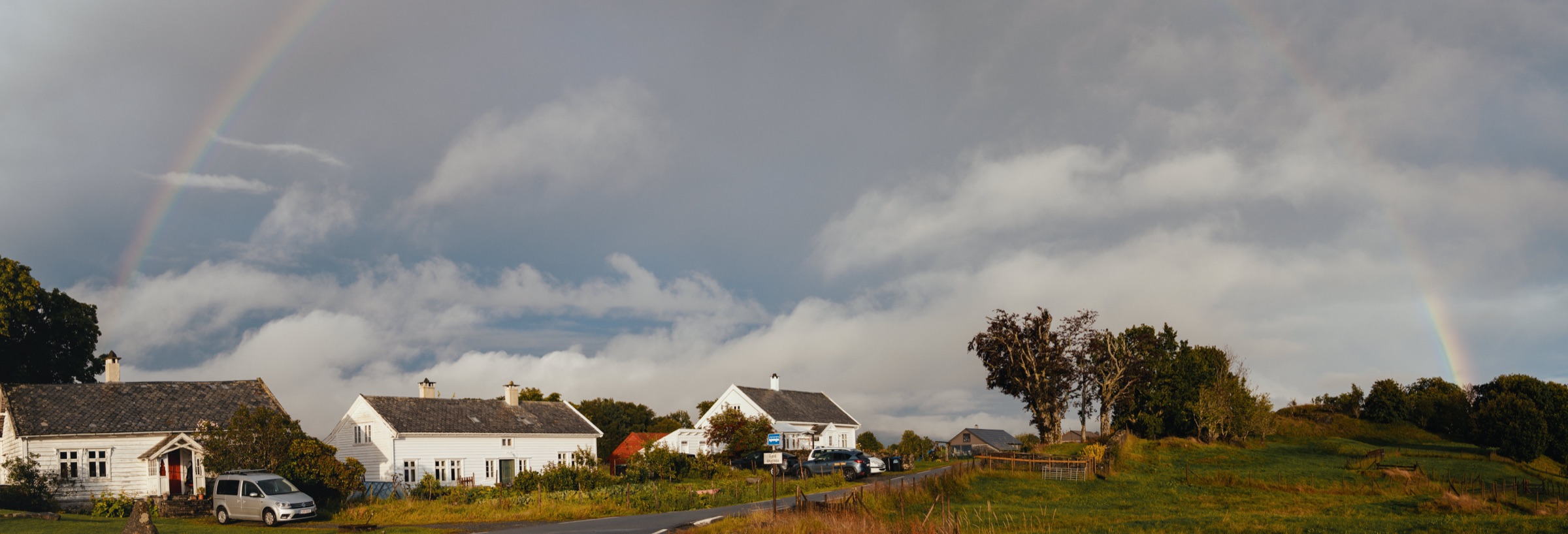 Sehr breites Panorama: einzelne Höfe in weiten Wiesen unter einem hohen, aufreißenden Wolkenhimmel.