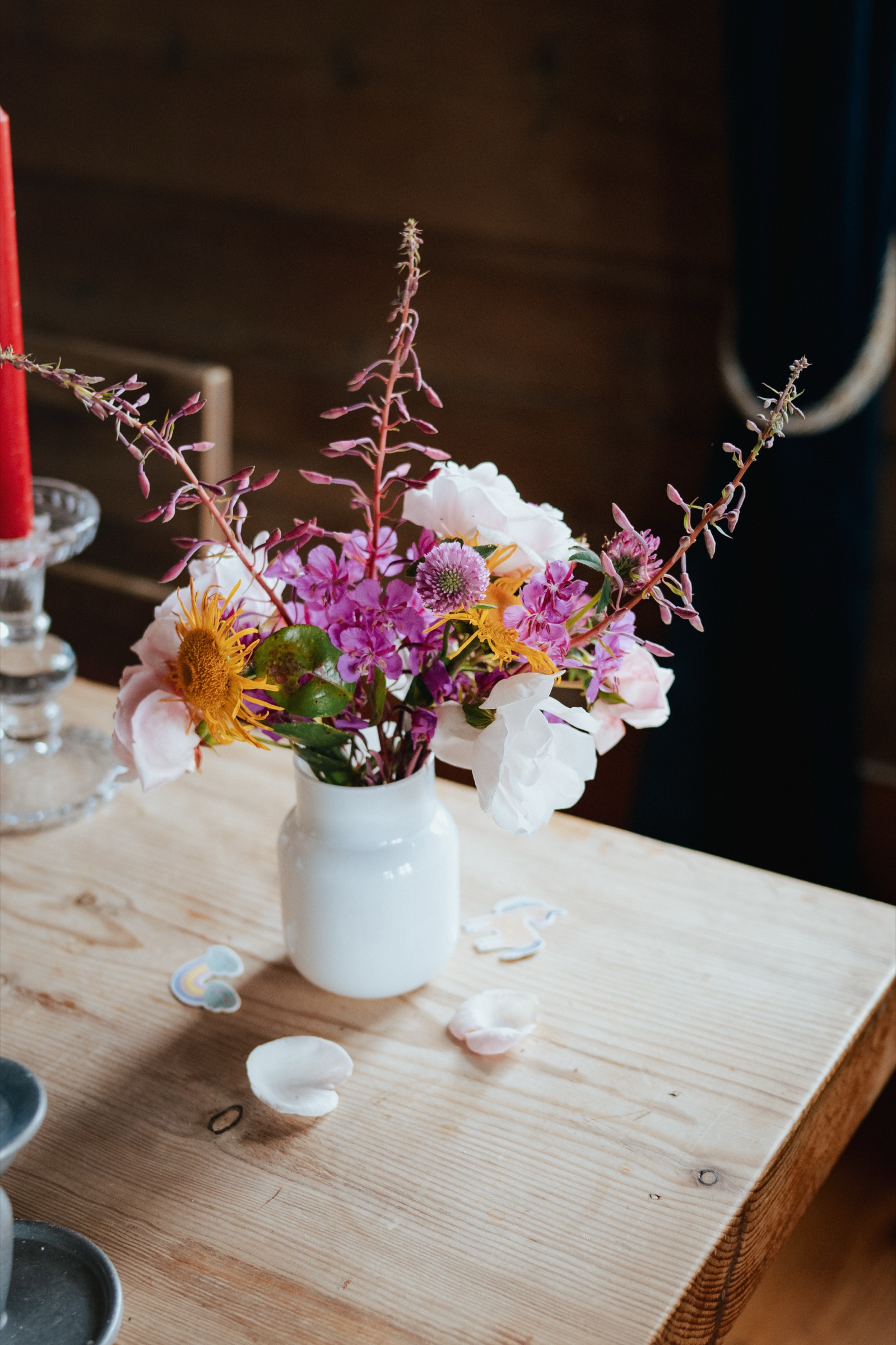 Hochformat: Wiesenblumen in einer weißen Kanne neben brennenden Kerzen auf einem Holztisch.